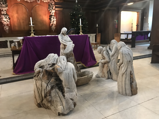 Krippe in einer Kirche mit Statuen, einem Tisch mit einer Decke und Kerzen, Stühlen, einem geschmückten Weihnachtsbaum und einer Wand im Hintergrund.