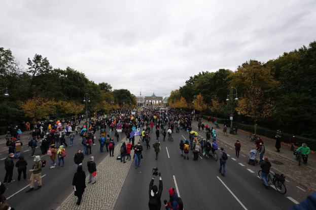 Eine große Gruppe von Menschen geht eine von Bäumen gesäumte Straße mit Laternenpfählen entlang und hält Kameras während einer Protestdemo in Berlin mit einem Gebäude und einem klaren Himmel im Hintergrund.
