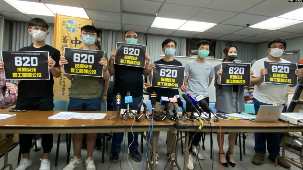 Eine Gruppe maskierter Personen mit Schildern versammelt sich um einen Tisch mit Mikrofonen, Papieren und Laptops und protestiert gegen das Auslieferungsgesetz in Hong Kong, mit einem Banner und beleuchteter Decke im Hintergrund.