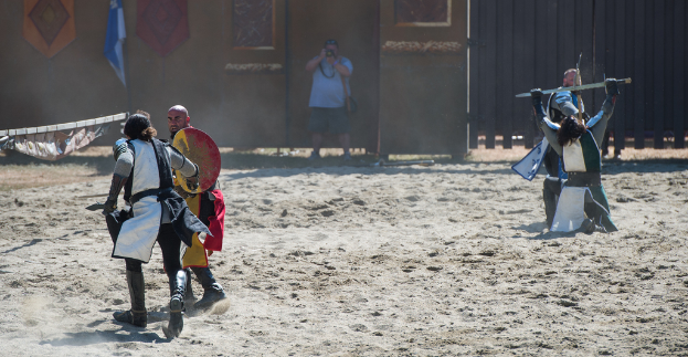 Eine Person steht in der Mitte und hält eine Kamera, während sie ein Bild von einem Kampf zwischen zwei Gruppen auf beiden Seiten aufnimmt, mit einer Wand, einem Tor und einer Flagge im Hintergrund und Sand unten.