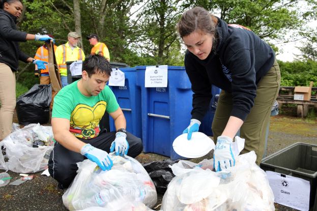 Eine Gruppe von Menschen sammelt Müll in einem Park, mit einem Mann und einer Frau in Handschuhen, die Abfall auf Tellern sammeln, umgeben von Plastik, Flaschen und Schutt, in der Nähe eines Mülleimers und einer Bank unter Bäumen mit einem klaren Himmel.