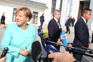 Bundeskanzlerin Angela Merkel spricht zu Reportern vor dem Kanzleramt in Berlin, umgeben von Menschen mit Mikrofonen und Gebäuden im Hintergrund.