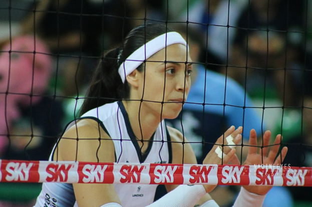 Eine weibliche Volleyballspielerin in Uniform springt in der Nähe des Netzes mit konzentrierter Miene, der Court und ein unscharfer Hintergrund dahinter.
