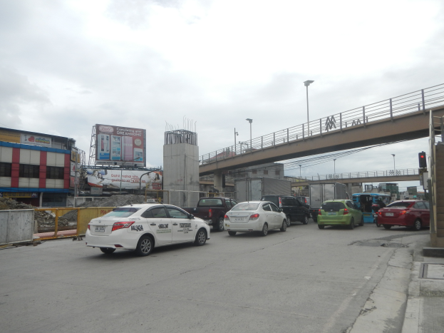 Eine belebte Straße mit parkenden Autos, eine Brücke mit Geländern und Laternen, Ampeln, Gebäude mit Fenstern, Schilder mit Text und Bildern und ein bewölkter Himmel im Hintergrund.