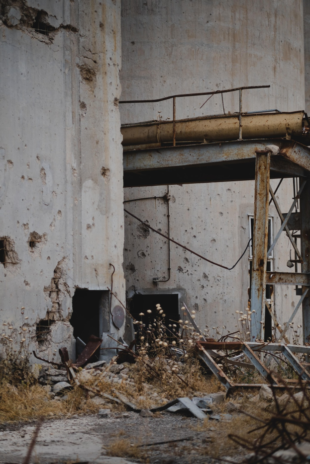 Altes Geb├Ąude mit einer zentralen Metallstruktur, umgeben von Pflanzen, Eisenstangen und Schutt, mit brüchigen W├Ąnden und zerbrochenen Fenstern.