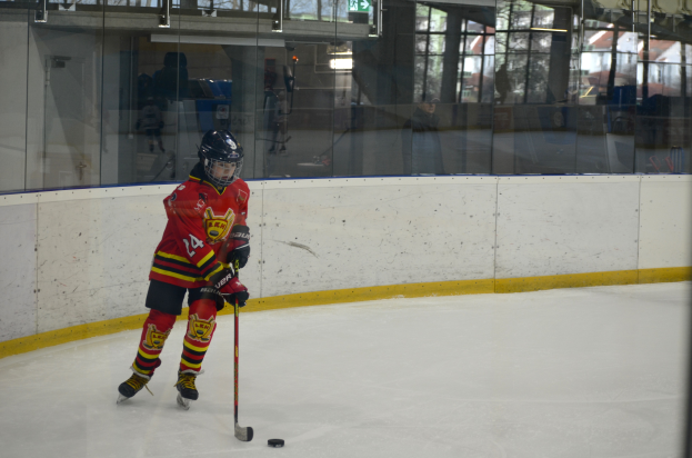 Ein junger Junge mit Helm und Hockeystock auf einem Eisstadion mit Glaswand spielt, umgeben von Menschen, Bäumen, Gebäuden und Himmel.