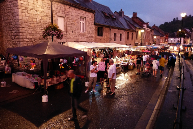 Eine Gruppe von Menschen auf einer Straße mit Geschäften, Schirmen, hängenden Spielzeugen, Taschen, Gebäuden mit Fenstern, Bäumen, Straßenlaternen und Barrieren.