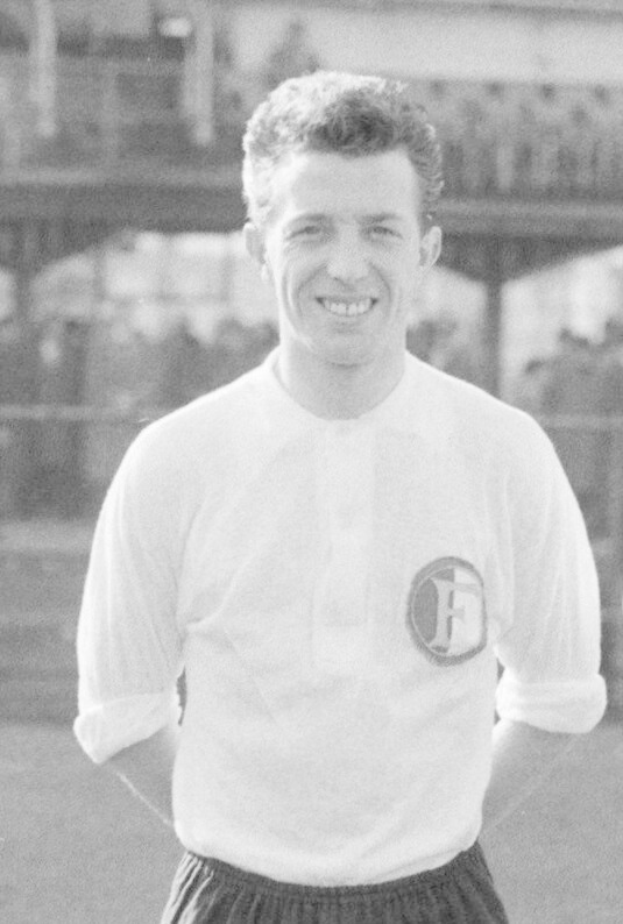 Ein Schwarz-Weiß-Foto eines grinsenden Mannes in T-Shirt und Shorts auf einem Fußballfeld mit einem Gebäude im Hintergrund, der als Mario Mandžukić identifiziert wird.
