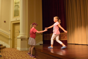 Zwei junge Mädchen tanzen auf einer Bühne vor einem Vorhang, mit einer Person, die links auf einem Stuhl sitzt, einer Säule und einer Wand im Hintergrund.