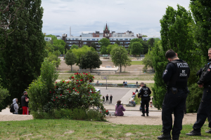 Zwei Polizisten stehen vor einer Gruppe von Menschen in einem Park mit grünem Gras, Bäumen, bunten Blumen, Gebäuden, Pfählen und einem klaren blauen Himmel.