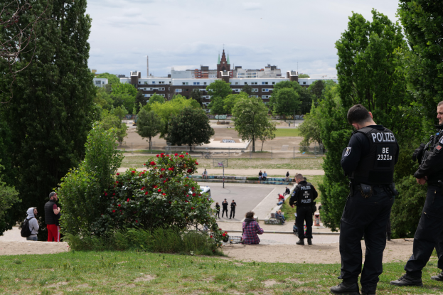 Zwei Polizisten stehen vor einer Gruppe von Menschen in einem Park mit grünem Gras, Bäumen, bunten Blumen, Gebäuden, Pfählen und einem klaren blauen Himmel.