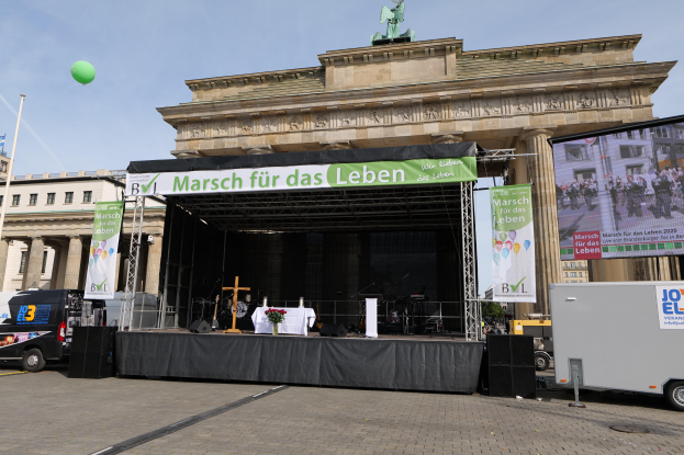 Bühne vor dem Brandenburger Tor mit einem Tisch, der eine Blumenvase, ein Kreuzsymbol und Banner enthält, begleitet von Lautsprechern und anderen Gegenständen, mit Fahrzeugen auf der Straße und Gebäuden im Hintergrund.