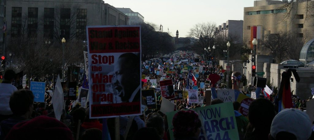 Menschen mit Plakaten auf einer Straße, im Hintergrund Gebäude, Bäume, Laternenpfähle und der Himmel.