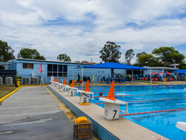 Ein großes Schwimmbad mit schwimmenden Menschen, Bahnmarkierungen, Verkehrskegeln, Stühlen, Sonnenschirmen, einem Gebäude mit Fenstern, einer Flagge, Bäumen und einem bewölkten Himmel im Hintergrund.
