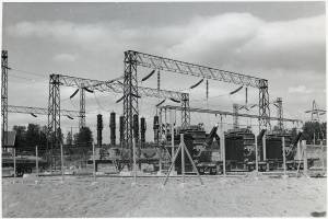 Ein Schwarz-Weiß-Foto eines elektrischen Umspannwerks mit Transformatoren, Polen, Drähten, Maschinen, einem Zaun, Sand, Bäumen und einem bewölkten Himmel, mit Text unten.