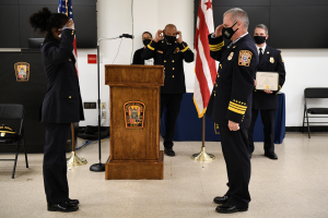 Gruppe uniformierter Personen in Masken vor einem Rednerpult während einer Vereidigung, eine hält ein Zertifikat, mit Flaggen und Sitzplätzen.