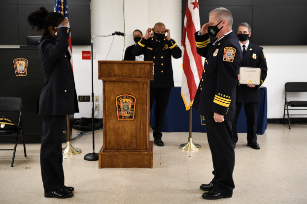 Gruppe uniformierter Personen in Masken vor einem Rednerpult während einer Vereidigung, eine hält ein Zertifikat, mit Flaggen und Sitzplätzen.