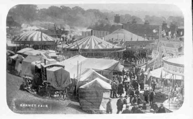 Ein altes Schwarz-Weiß-Foto einer belebten KarnevalsSzene mit Zelten, Menschen, Wagen, Pfählen, Bäumen und Gebäuden im Hintergrund, beschriftet mit "Barnet Fair, London, England".