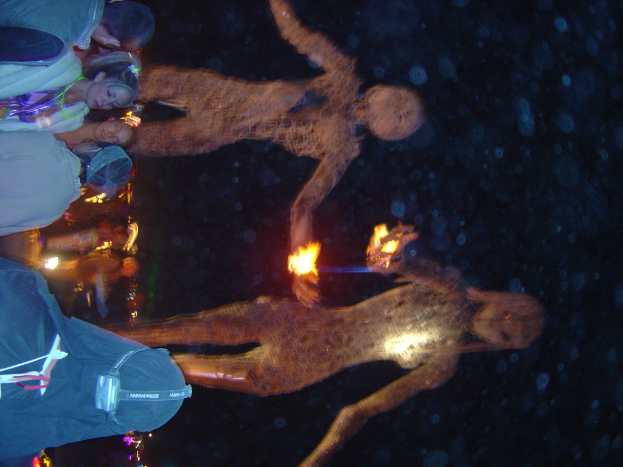 Mehrere Personen stehen auf der linken Seite des Bildes, mit brennenden Statuen dahinter und Lichtern im Hintergrund.