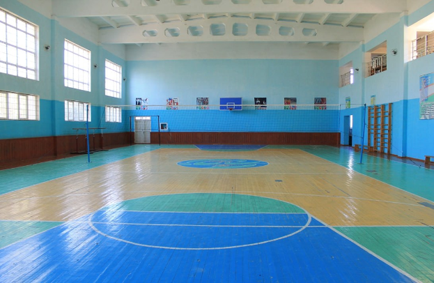 Großer Turnsaal mit einem blauen und weißen Basketballfeld, einem Netz an Stangen, einer Tür, Fenstern, Plakaten an der Wand, einem Geländer und einem Dach.