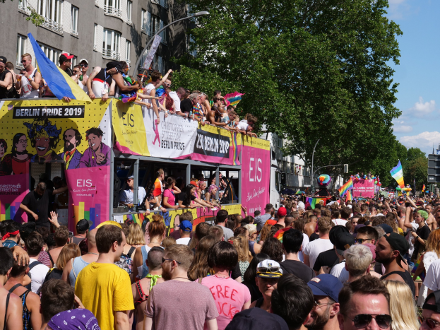 Eine große Menschenmenge marschiert auf der Straße bei der Christopher Street Day Parade in Berlin 2019, viele tragen Mützen und Schutzbrillen, halten Fahnen, mit Bannern und Gebäuden im Hintergrund bei bewölktem Himmel.