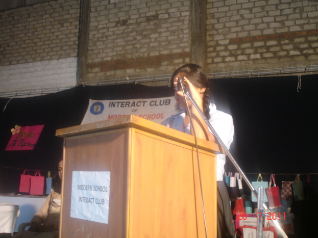 Ein Mädchen in einem weißen Hemd steht neben einem Rednerpult in einem Konferenzraum, mit einem Mikrofonständer rechts daneben, einem weißen Banner im Hintergrund und Taschen, die an einem Faden rechts hängen.