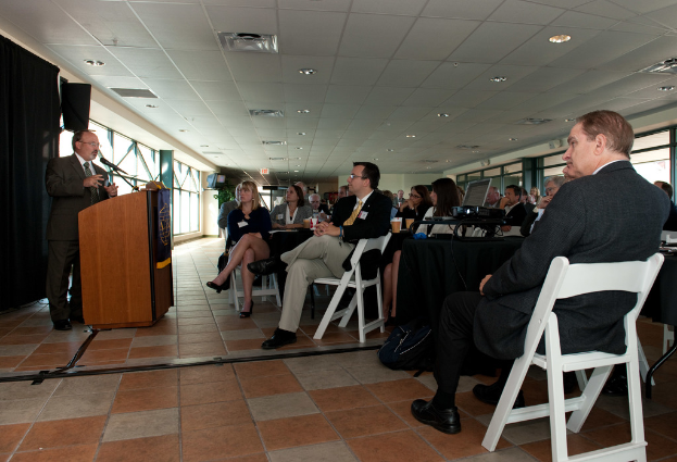 Eine Gruppe von Menschen, darunter Männer und Frauen, sitzt einem Mann gegenüber, der an einem Rednerpult steht und in ein Mikrofon spricht, mit Fenstern im Hintergrund.
