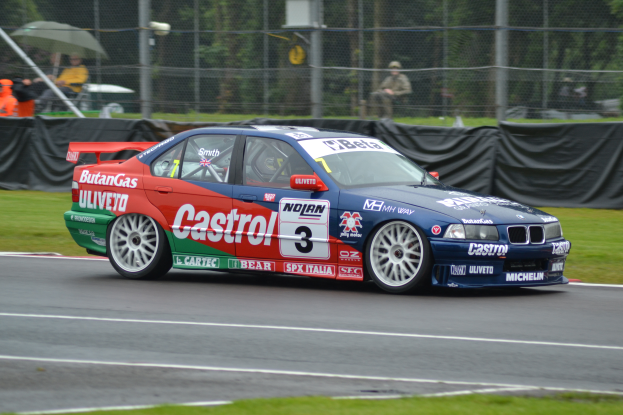 Ein Auto rennt auf einer Rennstrecke mit Zuschauern hinter einem Maschendrahtzaun und Bäumen im Hintergrund.