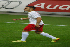 Ein Mann in einem weißen Hemd und roten Shorts, der Fußballschuhe trägt, läuft auf einem Fußballfeld mit einer Tafel im Hintergrund.