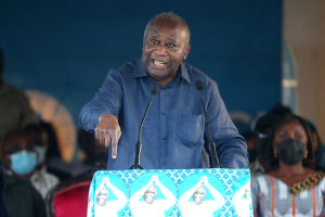 Ein Mann in einem blauen Hemd spricht an einem Pult mit zwei Mikrofonen, gegenüber einer sitzenden Menge, einige tragen Masken, mit einer Wand im Hintergrund.