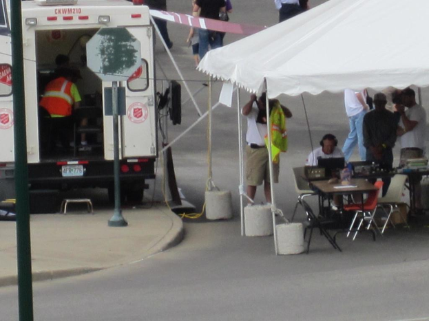 Ein Rettungswagen auf der linken Straßenseite, während auf der rechten Seite ein Zelt eine Gruppe von Menschen schützt, mit einem Tisch und einem Stuhl in der Nähe.