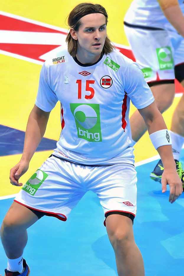 Ein Mann in weißer Uniform spielt Handball, mit zwei Personen im Hintergrund auf einem bunten Boden, wahrscheinlich bei der Handball-Weltmeisterschaft 2018.