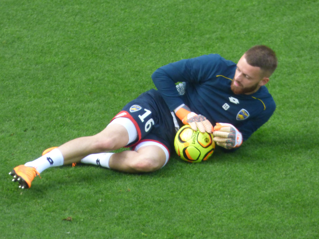 Ein Fußballspieler liegt auf dem Boden und hält einen Ball, trägt ein blaues T-Shirt, Shorts, Socken und Schuhe.