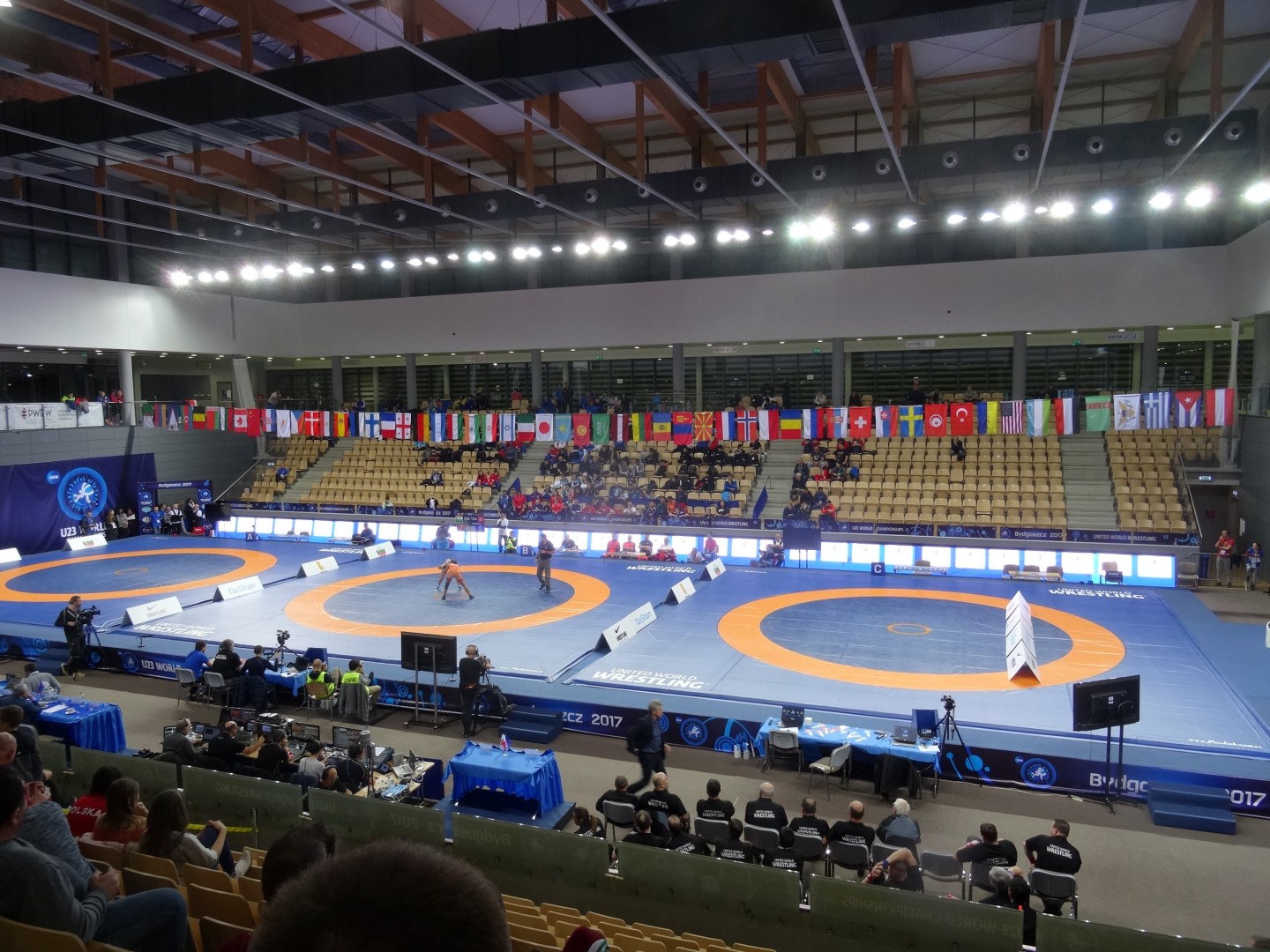 Großer Innenstadionsaal mit Zuschauern bei einem Wrestling-Kampf, mit Sitzplätzen und Stehplätzen, Tischen mit Gegenständen und Hintergrundflaggen, Lichtern und Stangen.