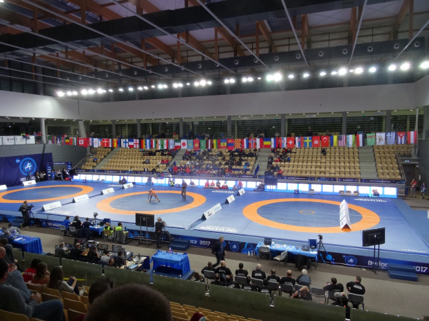 Großer Innenstadionsaal mit Zuschauern bei einem Wrestling-Kampf, mit Sitzplätzen und Stehplätzen, Tischen mit Gegenständen und Hintergrundflaggen, Lichtern und Stangen.