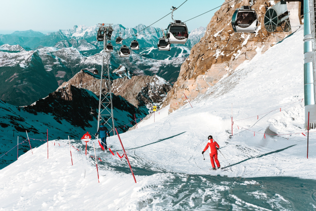 Gruppe von Menschen, die eine schneebedeckte Piste mit einem Seilbahnwagen im Hintergrund hinabskien, unter einem klaren blauen Himmel mit einer Gebirgskette.