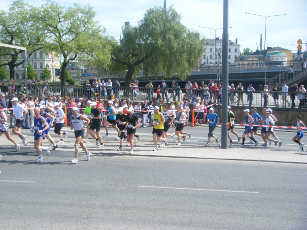 Gruppe von Läufern bei einem Marathon auf einer Straße mit Zielpfosten und Band, umgeben von Metallzaun und Barrieren, mit Zuschauern auf dem Gehweg, Pfosten, Schilder, Brücke, Gebäuden, Bäumen und bewölktem Himmel.