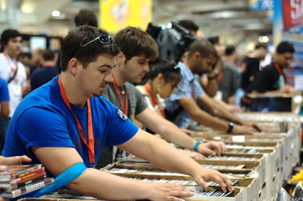 Eine Gruppe von Menschen mit Ausweisen packt Bücher in Kisten, während über ihnen Lampen leuchten und ein Banner auf der rechten oberen Seite zu sehen ist, während einige andere im Hintergrund stehen.