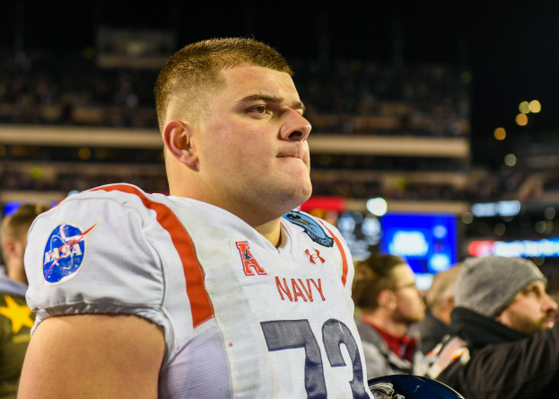 Ein Mann in einer marineblauen Uniform steht auf einem Footballfeld und hält einen Helm in der Hand, mit unscharfen Menschen, Lichtern und einem Bildschirm im Hintergrund.