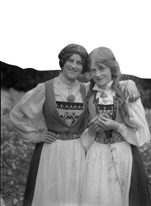 Zwei junge Frauen in traditioneller bayrischer Tracht, mit weißen und schwarzen Kleidern mit aufwendiger Stickerei und Perlen, posieren vor der Kamera gegen einen Hintergrund aus üppigen grünen Pflanzen und einem hellen weißen Himmel.