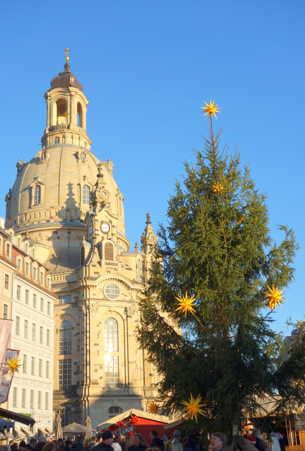 Ein geschäftiger Weihnachtsmarkt in Dresden, Deutschland, mit einem großen Weihnachtsbaum vor einer Kirche, vielen Menschen drumherum und einem Banner mit Text auf der linken Seite, unter einem sichtbaren Himmel.