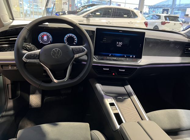 Innenansicht eines Volkswagen e-Golf mit Lenkrad, Armaturenbrett, Bildschirmen, Knöpfen, Sitzen und Glastüren, mit anderen Autos, Gebäuden, Bäumen, Pfählen und Himmel im Freien.