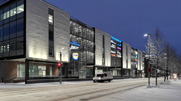 Ein Lieferwagen fährt auf einer verschneiten Straße nachts, umgeben von Gebäuden, Straßenlaternen, Verkehrspfosten, Verkehrszeichen, Schildern, Bäumen und einem Sternenhimmel.