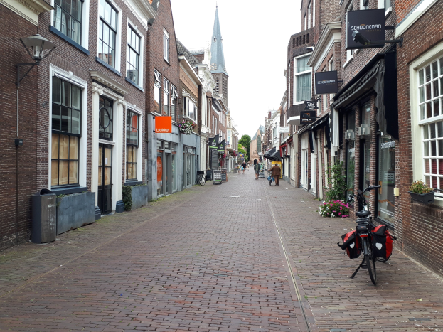 Ein Fahrrad, das auf einer städtischen Straße in Delft, Niederlande, geparkt ist und von Gebäuden mit Fenstern, Blumentöpfen und Informationsschildern flankiert wird, mit Bäumen und einem klaren Himmel im Hintergrund.