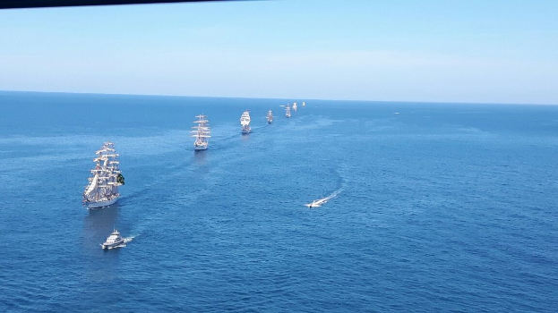 Eine große Gruppe von Segelschiffen auf dem Meer während eines Regattas, unter einem klaren blauen Himmel mit helllem Sonnenlicht, das sich auf dem Wasser spiegelt.