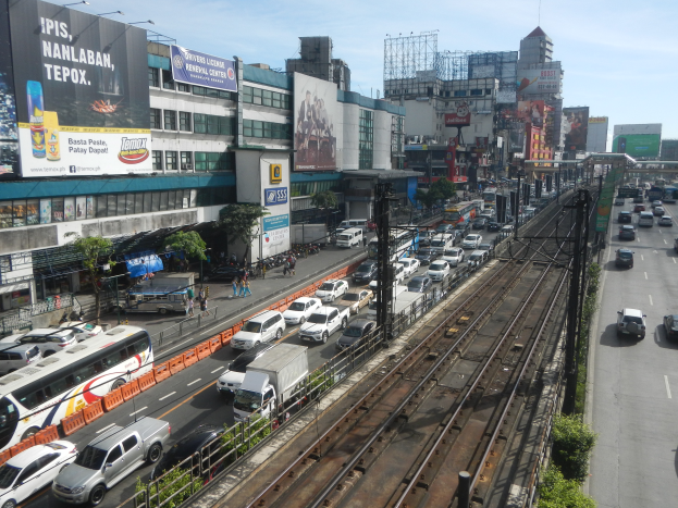 Eine belebte Stadtstraße mit starkem Verkehr, Fußgängern, hohen Gebäuden und einem bewölkten Himmel im Hintergrund.