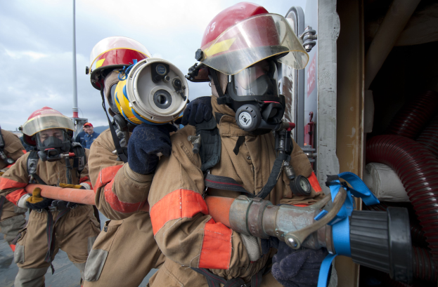 Feuerwehrleute in Schutzausrüstung mit einem, der einen Schlauch hält, einem Pfahl im Hintergrund, einem bewölkten Himmel und Rohren auf der rechten Seite.