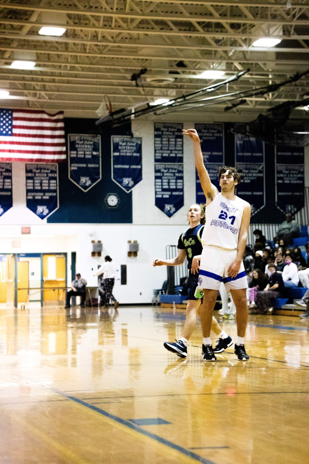 Ein Mann in Sportbekleidung führt einen Basketballwurf in einer Turnhalle aus, während Zuschauer von den Tribünen zusehen.