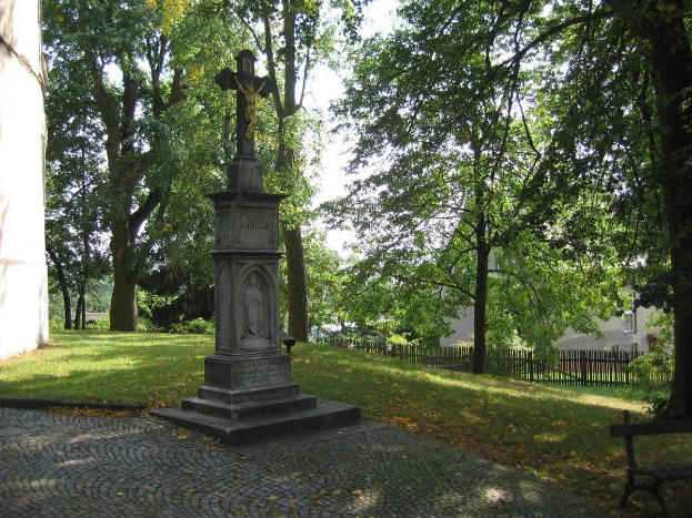 Ein Denkmal steht in einem Park mit einer Bank auf der rechten Seite, umgeben von Gras, Bäumen und einem Gebäude im Hintergrund unter einem klaren Himmel.