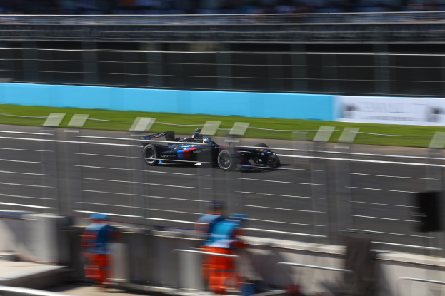 Ein Formel-1-Auto fährt auf einer Rennstrecke an einer Menge von Zuschauern vorbei, Gras auf dem Boden und Schilder mit Text im Hintergrund.